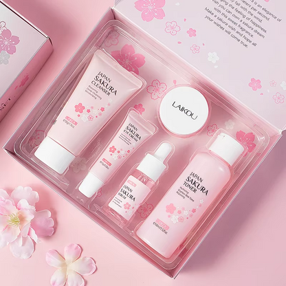 Lensemble de soins du visage Laikou Sakura de Jay Cosmetic est présenté sur une surface rose avec son Nettoyant Visage Sakura, son toner, sa crème, son sérum à la niacinamide et son baume à lèvres dans un emballage sur le thème des fleurs de cerisier. Une boîte ouverte révélant un motif floral se trouve au milieu de pétales de fleurs de cerisier dispersés.