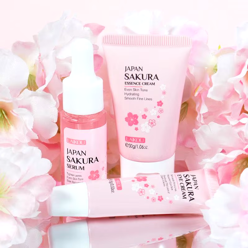 Lensemble de soins du visage Laikou Sakura de Jay Cosmetic est présenté sur une surface rose avec son Nettoyant Visage Sakura, son toner, sa crème, son sérum à la niacinamide et son baume à lèvres dans un emballage sur le thème des fleurs de cerisier. Une boîte ouverte révélant un motif floral se trouve au milieu de pétales de fleurs de cerisier dispersés.