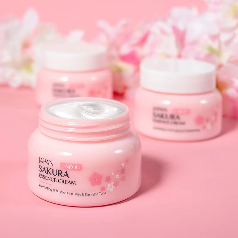 Lensemble de soins du visage Laikou Sakura de Jay Cosmetic est présenté sur une surface rose avec son Nettoyant Visage Sakura, son toner, sa crème, son sérum à la niacinamide et son baume à lèvres dans un emballage sur le thème des fleurs de cerisier. Une boîte ouverte révélant un motif floral se trouve au milieu de pétales de fleurs de cerisier dispersés.